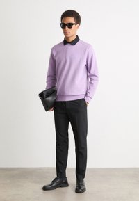 Λιλά πουλόβερ με μαύρο γιακά, υφασμάτινη υφή, λεπτομέρεια μπροστινής τσέπης; σε σύμπραξη με μαύρο παντελόνι και παπούτσια. Κρατώντας μια μαύρη τσάντα clutch.