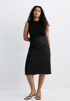 Mujer con cabello largo y rizado, vestida con un vestido negro sin mangas y sandalias negras de tacón con tiras, de pie frente a un fondo blanco liso.