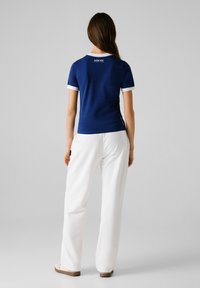 Donna in piedi di spalle, che indossa una maglietta blu navy con polsini bianchi e pantaloni bianchi, su uno sfondo chiaro e semplice.