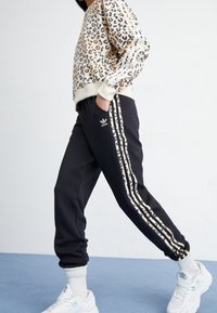 Felpa leopardata beige, abbinata a pantaloni della tuta neri con strisce bianche ai lati e dettagli neri. Scarpe da ginnastica bianche.