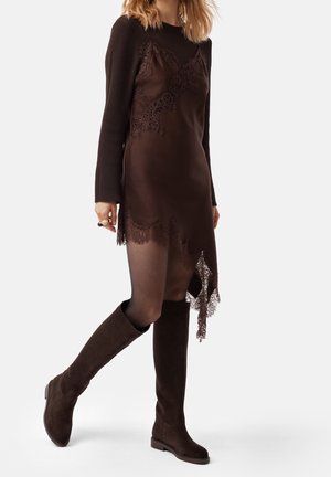 Nainen kävelee ruskeaa pitsireunusta olevaa liivimekkoa, ribbineulosta pitkähihainen toppia, läpikuultavia sukkahousuja ja polven korkeudella olevia mokkanahkasaappaita käyttäen.