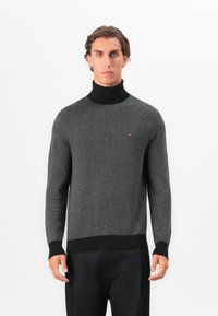 Hombre con suéter de espiga en gris y negro, con cuello de tortuga negro y pantalones negros, de pie frente a un fondo blanco liso.