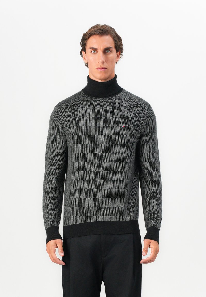 Hombre con suéter de espiga en gris y negro, con cuello de tortuga negro y pantalones negros, de pie frente a un fondo blanco liso.