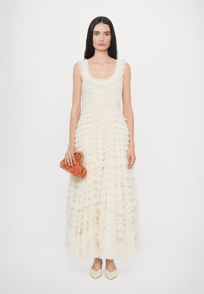 Witte, mouwloze jurk met gelaagde ruffles; gestructureerde stof. Gecombineerd met een oranje kralen clutch en crèmekleurige puntige schoenen.