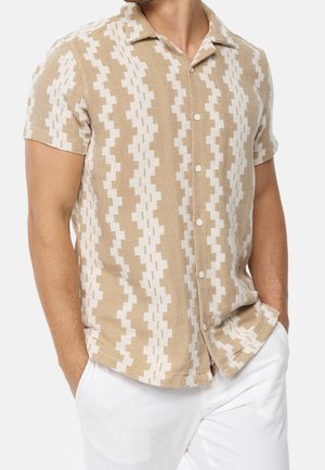 Man wearing short-sleeve beige button-up shirt with white zigzag pattern, paired with white pants, hands in pockets.