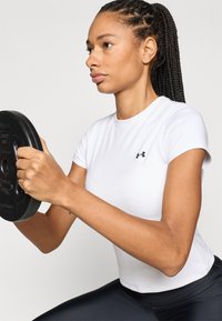 Bílá atletická košile s krátkým rukávem a černým logem, doplněná černými přiléhavými kalhotami. Osoba drží kulatou černou zátěžovou destičku.