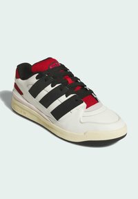 White athletic shoe with black stripes, textured red mesh tongue, and black laces. Features a light gray sole and iconic branding.