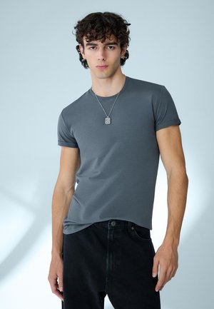 Mladý muž s kudrnatými tmavými vlasy, oblečený v těsně padnoucím šedém tričku, černých kalhotách a se stříbrným přívěskem na krku, stojící před jednobarevným pozadím.