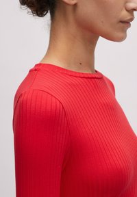 Rotes geripptes Langarmoberteil mit Rundhalskragen; der Stoff zeigt vertikale Streifen und eine glatte Textur, enganliegende Silhouette, die die Form betont.