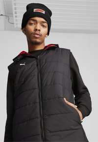 Czarna ocieplana kamizelka z czerwonymi akcentami, zapinana na zamek z przodu, z fakturowanej tkaniny, z logo Puma na piersi. Noszona na czarnej koszuli z długim rękawem i wełnianej czapce.