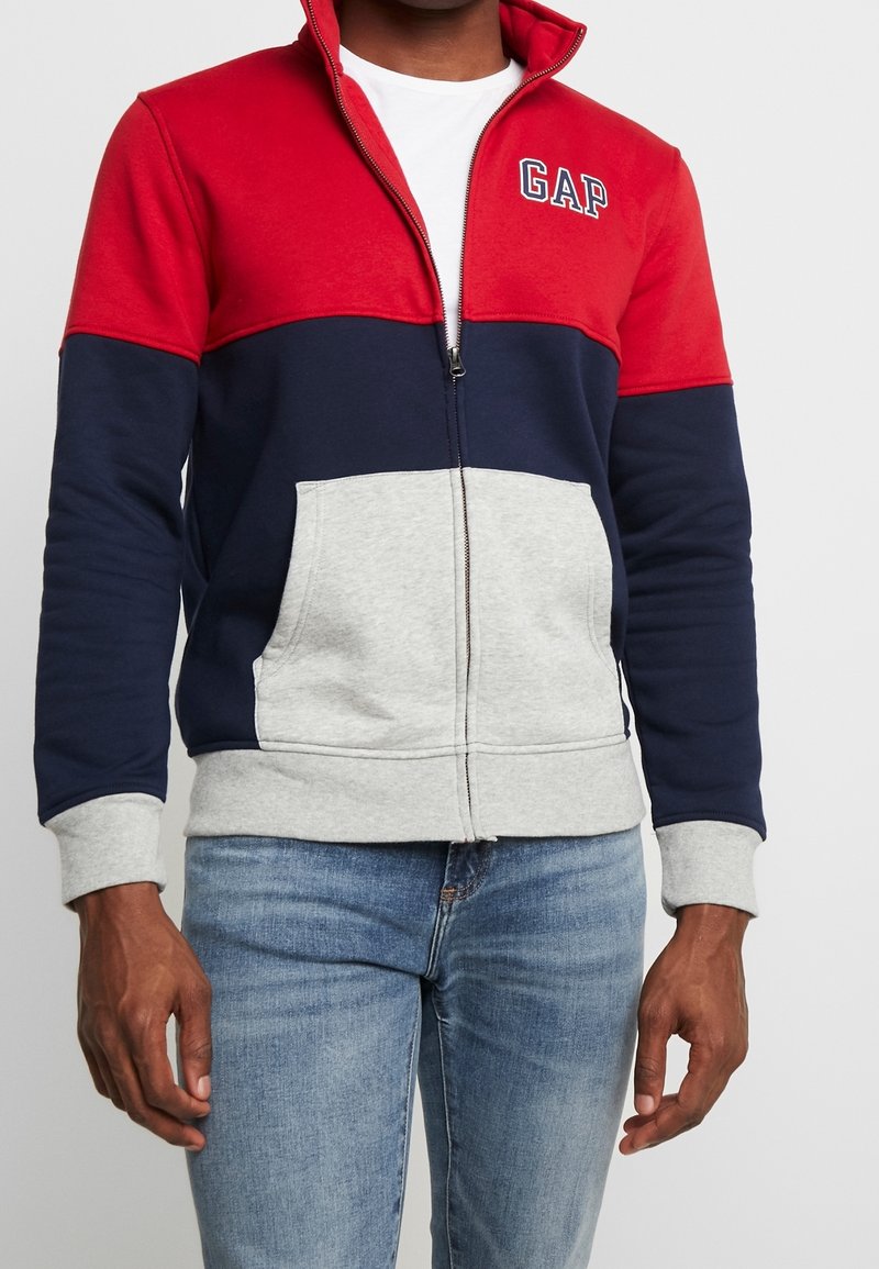 Homme portant une veste zippée rouge, bleu marine et grise avec le logo GAP sur la poitrine et un jean bleu clair, debout devant un fond uni.