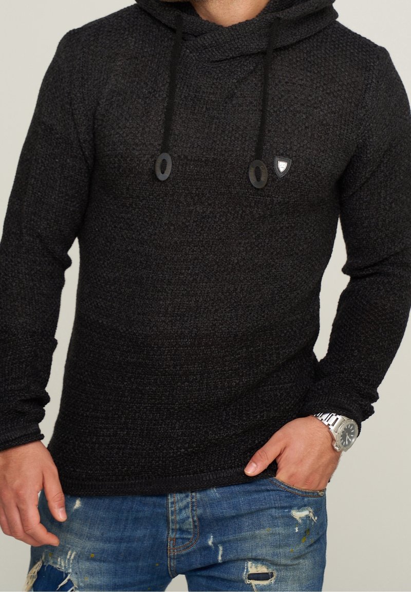 Sweat à capuche noir texturé avec une coupe ample, des cordons de serrage et un petit logo. Porté avec un jean bleu déchiré et une montre argentée.