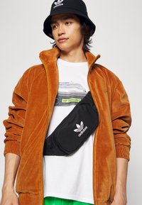 Ung person som bär en svart Adidas bucket hat, brun zip-up jacka, vitt grafiskt t-shirt och svart Adidas crossbody-väska.