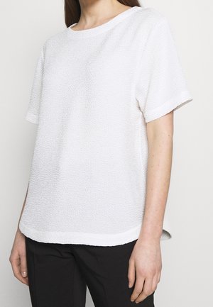 White short-sleeve top made of textured material, with a round neck and side slits at the hem, styled with black pants.