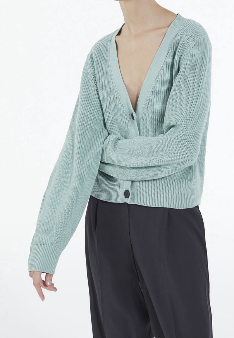 Ljusgrön stickad kofta med djup V-ringning, lång ärm och två knappar framtill. Matchas med mörka byxor.