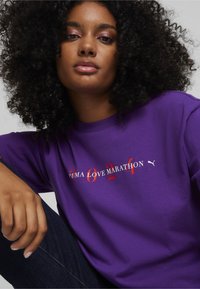 Violet trumpas rankovės marškinėliai su „PUMA LOVE MARATHON 2024“ tekstu raudona ir balta spalvomis, su mažu baltu PUMA logotipo akcentu. Minkšta medžiaga.
