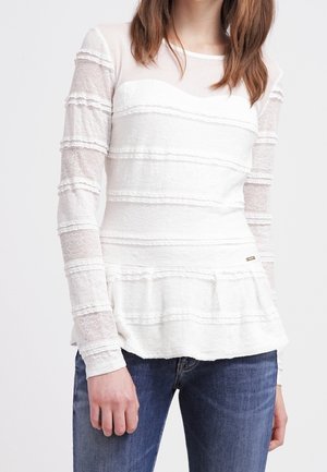 Kobieta ubrana w biały, długi rękaw, teksturowaną koronkową bluzkę w poziome paski z falbaną na dole, w połączeniu z niebieskimi dżinsami.