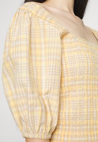 Blusa amarela com padrão xadrez, mangas bufantes, corpete drapeado e tecido texturizado com um aspeto leve e arejado.
