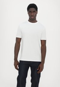 Hvit bomull T-skjorte med korte ermer og rund hals, med minimalistisk logo detaljer på brystet. Båret med mørk denim jeans.