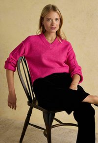 Pulover roz aprins, tricotat gros, cu decolteu în V și mâneci rulate, purtat cu pantaloni de catifea negri, așezat pe un scaun din lemn, pe un fundal neutru.