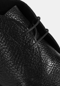 Schwarzer Lederschuh mit strukturierter Oberfläche, sichtbaren Nähten, zwei Schnürsenkelösen und einem glatten Finish an der Zunge.