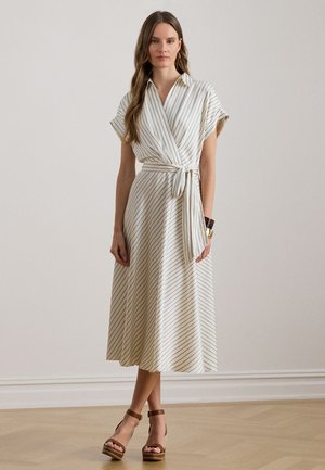 Donna che indossa un abito avvolgente color crema con sottili strisce blu e cintura annodata in vita, abbinato a sandali platform marroni, in piedi su un pavimento in legno.