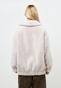 Giacca in finta pelliccia di un beige chiaro con colletto ampio, spalle ribassate e orlo elasticizzato. Presenta una texture liscia e dettagli con zip.
