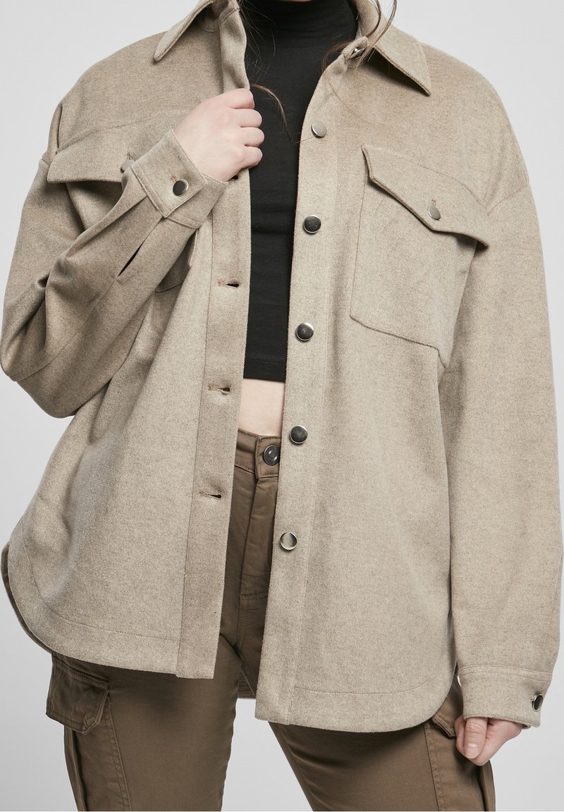 Béžová vlněná košile-kabát s límečkem, knoflíkovým zapínáním a dvěma předními kapsami. Má uvolněný střih a strukturovanou tkaninu.