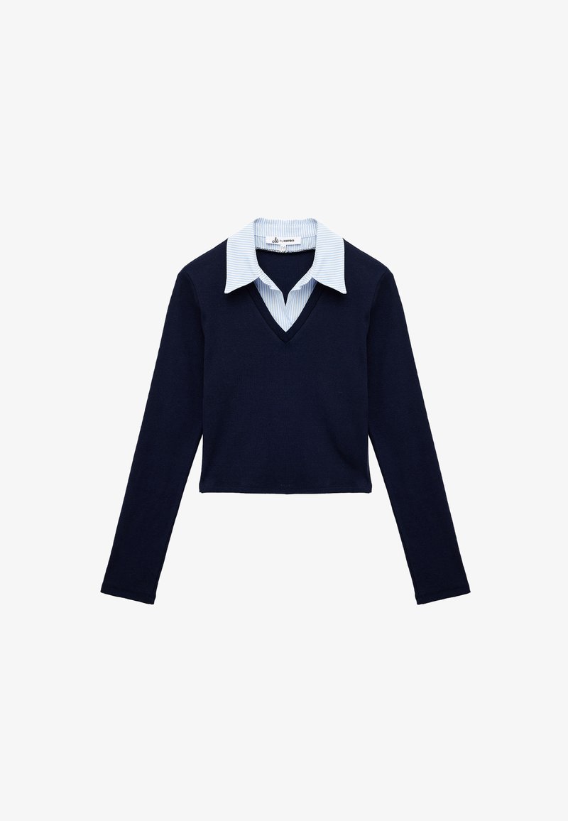 Granatowy prążkowany sweter o krótkim kroju z dekoltem w kształcie litery V, w zestawieniu z jasnoniebieską i białą koszulą w paski z kołnierzykiem na dole.