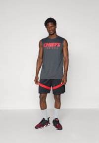 Pilkas be rankovių marškinėliai su raudonu užrašu "CHIEFS FOOTBALL", derinami su juodomis šortais su raudonais akcentais ir pilkais kojinėmis. Juodi ir raudoni sportbačiai.