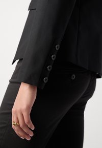 Gros plan d'une personne portant un blazer noir et un pantalon, montrant les boutons de manche et une bague en or à la main gauche.