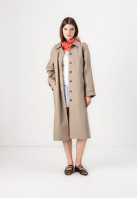 Beige Trenchcoat mit Knöpfen, lässiger Schnitt und weiten Ärmeln. Getragen über einem weißen Oberteil und Jeansshorts, ergänzt durch einen roten Schal.