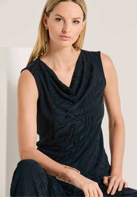 Mouwloos zwart top met een gedrapeerde col en een textuurpatroon van golven. Bevat glanzende accenten en is gecombineerd met metallic sieraden.