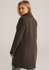 Il cappotto di lana marrone presenta una vestibilità sartoriale, colletto a rever e polsini bottonati. Il retro mostra un taglio pulito con cuciture verticali.