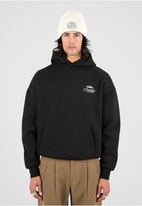 Sudadera de algodón negra con bolsillo canguro y logotipo bordado. Combinada con pantalones beiges con pliegues y un gorro de punto crema con un parche.
