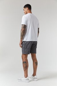 Weißes Baumwoll-T-Shirt und graue Cargo-Shorts mit Seitentaschen, kombiniert mit weißen Sportschuhen. Auffällige Tattoos an den Beinen sichtbar.