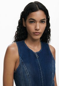Denim dress with a sleeveless design, featuring a round neckline, front zipper, and contrasting stitching in a dark blue shade.