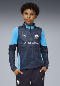 Felpa atletica blu e nera con motivo testurizzato, zip a metà, dettagli con logo e design strutturato. Indossata con pantaloni scuri.