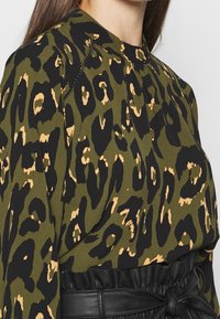 Blusa verde con stampa leopardata e accenti neri, caratterizzata da un collo alto, tessuto texturizzato e un design plissettato sulle spalle.