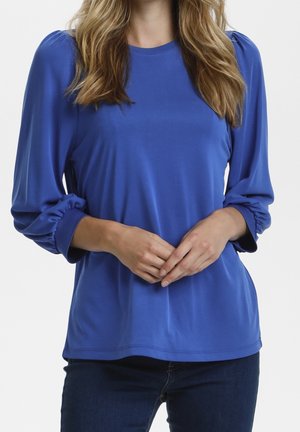 Femme portant un chemisier bleu roi à manches longues avec des poignets froncés, mains lâchement jointes devant, associé à un jean bleu foncé.