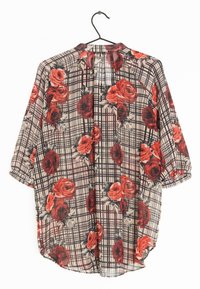 Blouse florale transparente avec des roses rouges et noires sur un motif à carreaux beige. Elle présente un col et des manches courtes, suspendue à un crochet noir.