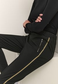 Pantalons noirs à la coupe ajustée, avec une rayure dorée sur le côté et une poche avec fermeture éclair. Fabriqués en tissu lisse avec une taille élastique.