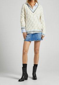Pull tricoté crème avec col en V, accents à rayures bleues, superposé sur une chemise à rayures bleu clair, associé à une mini jupe en denim et des bottines noires.