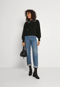 Fekete pulóver gyöngyökkel díszített gallérral, világoskék felhajtott szárú farmerrel és fekete, magas sarkú bokacsizmával párosítva. Fekete kézitáskát tart.