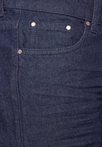 Tummansiniset farkut, joissa on klassinen muotoilu, näkyvät ompeleet, kuparin niitit ja sileä tekstuuri, korostaen etutaskua.