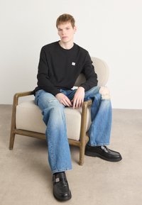 Zwart langemouwshirt met een klein logo, blauwe versleten jeans en zwarte schoenen met een dikke zool, zittend op een beige fauteuil.