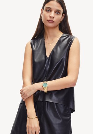 Zwart leren mouwloos topje met een V-hals en een ontspannen pasvorm. Model draagt een gouden horloge met een turquoise wijzerplaat en bijpassende armband.
