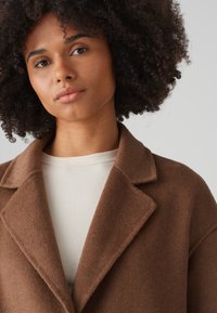 Brown wool coat with notch lapels, layered over a textured cream ribbed top. The coat features a soft, fuzzy texture and relaxed fit.