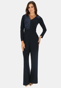 Granatowy kombinezon z długimi rękawami, akcentem z koronkowego wzoru kwiatowego na klatce piersiowej, dopasowany w talii i z szerokimi nogawkami, wykonany z gładkiego materiału.