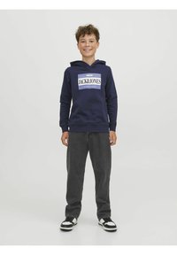 Sudadera con capucha azul marino y logo frontal, combinada con jeans negros y zapatillas en blanco y negro, de pie contra un fondo liso.